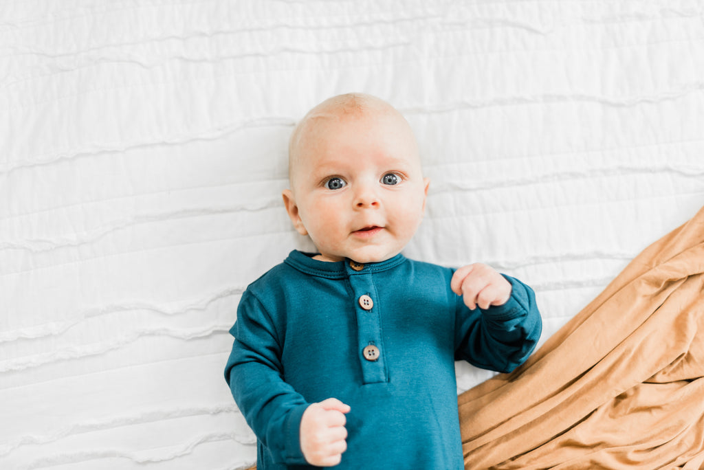 Baby boy in blue Kai romper laying on white bedding with camel Copper Pearl blanket